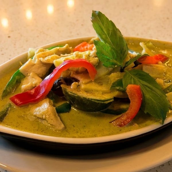 Green curry with chicken, red bell peppers, and basil in a white bowl on a speckled table.